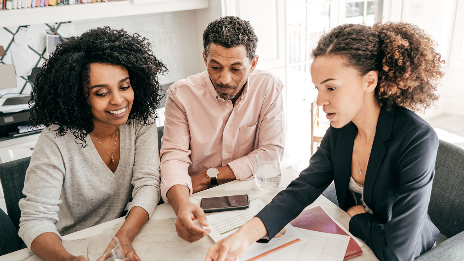 A couple is sitting at a table with an advisor. The advisor is gesturing toward a document, perhaps financial paperwork. The couple appears to be listening intently. The woman in the couple is smiling.