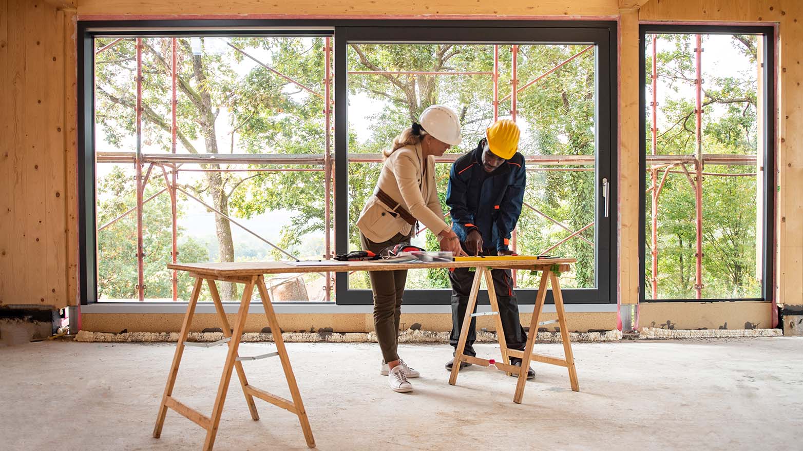 Two hard-hatted individuals are deep in discussion over plans, huddled around a table within the skeleton of a building in progress. One, a woman with flowing blonde locks, is smartly dressed in a beige blazer and khakis. The other, a Black figure, is donned in a navy jacket with a pop of orange, surveying the documents with equal intensity. The space is raw, with only the bare bones of the structure and the promise of what's to come visible through the expansive windows. The tableau is one of potential, of the excitement that comes with creation.