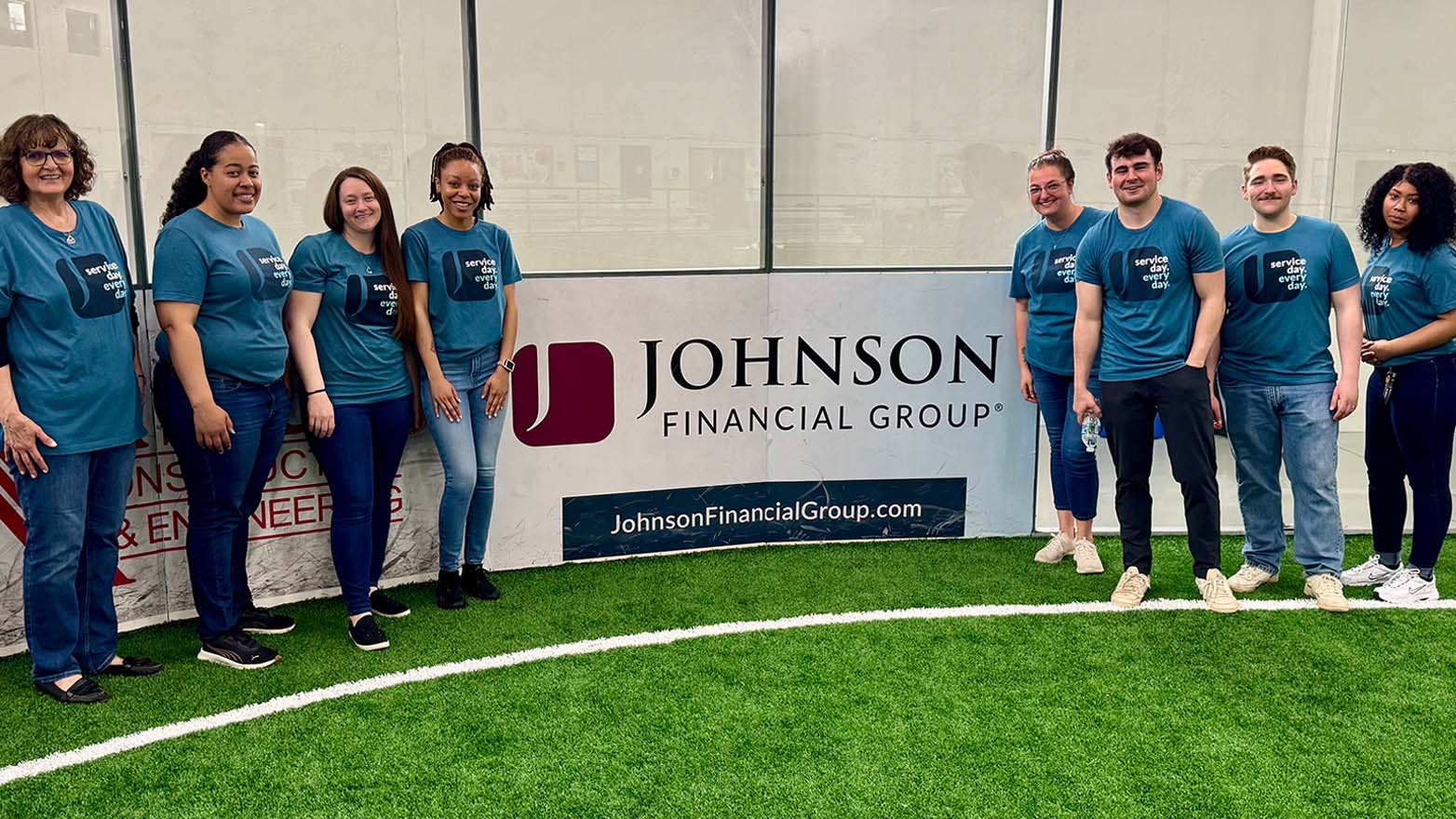 A group of people, wearing matching teal shirts with the slogan "Service day. Every day.", stand together in front of a Johnson Financial Group sign. The sign is set against a background of green turf with white lines.