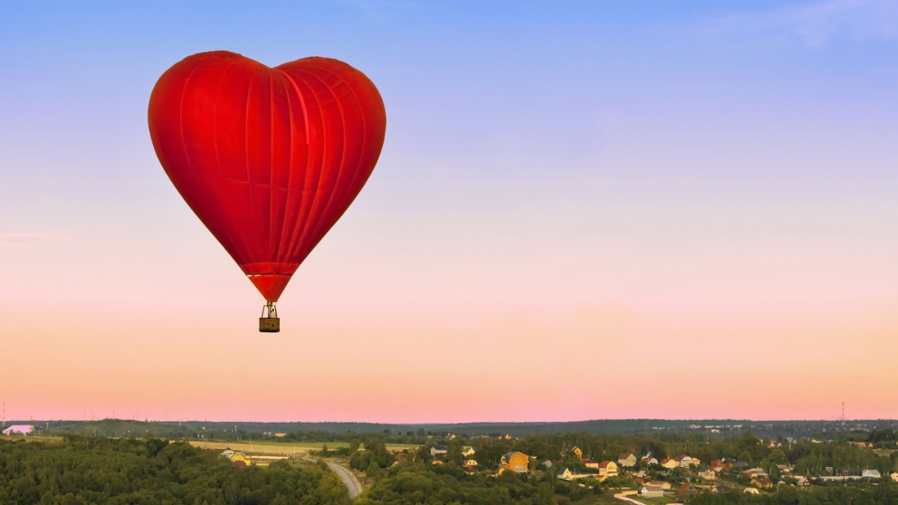 A red, heart-shaped hot air balloon soars above a serene landscape featuring a distant town and rolling green fields, set against a vibrant gradient sky.
