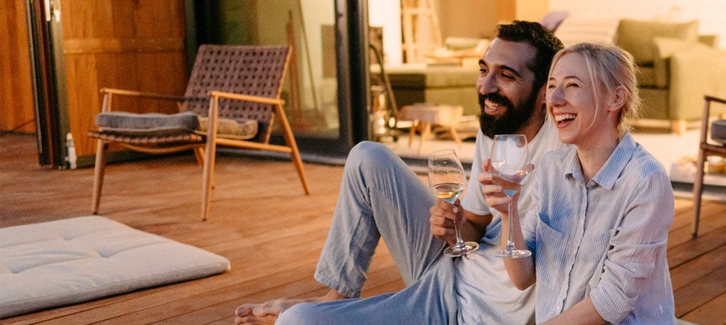 A bearded man and a blond woman are sitting on a wooden deck, holding wine glasses and laughing. They are barefoot and casually dressed. A cushioned mat sits beside them, and a wicker chair is visible in the background. Glass doors lead into the interior of the house.