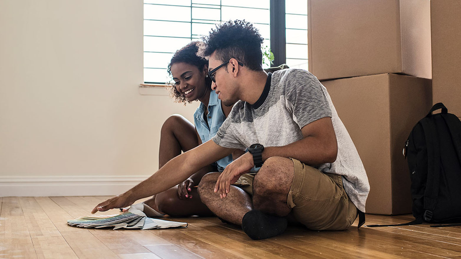 A young couple sits on the floor of their new home, contemplating paint swatches. The room is a jumble of moving boxes and a lone backpack. The woman grins as the man gestures at the color options. A window peeks out in the background.