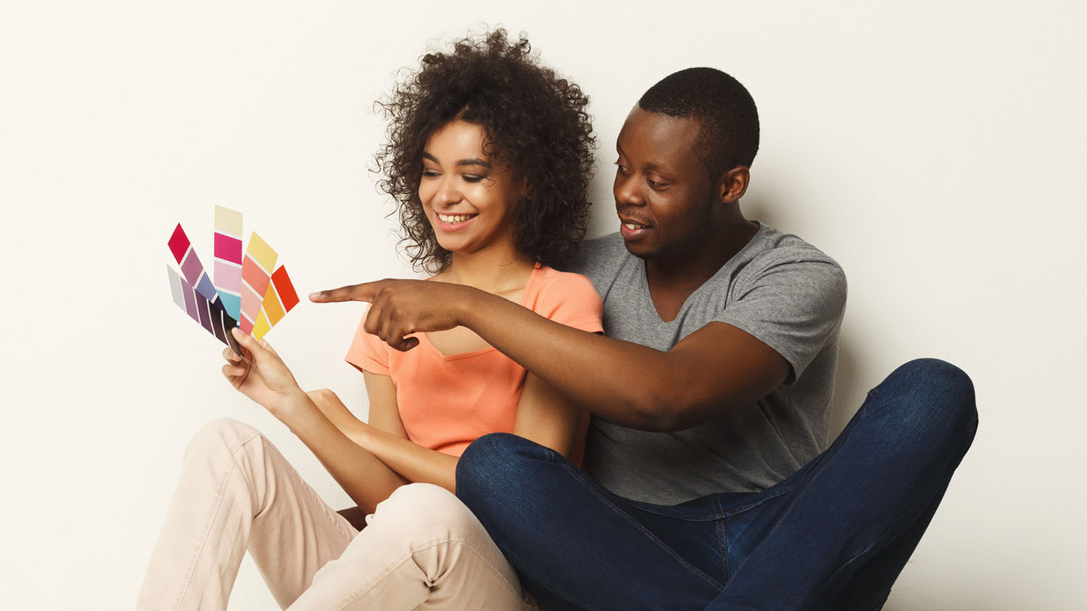 A smiling African American couple sitting on the floor in front of a white wall, reviewing paint color swatches. The man is pointing to a color on the swatches.