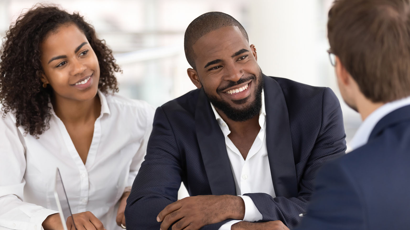 A smiling African American couple is engaged in a business meeting with a person who is out of focus and facing away from the camera. The couple is dressed professionally, with the woman in a white blouse and the man in a navy suit. The atmosphere suggests a positive and collaborative discussion.