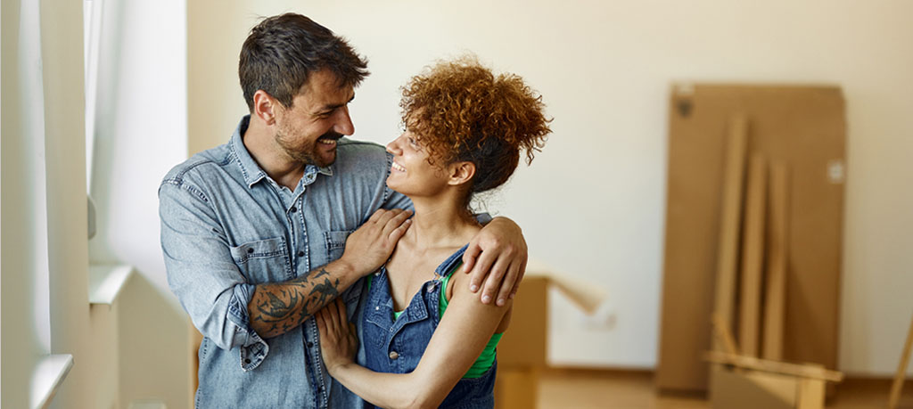 A young couple, bathed in the glow of a harshly bright room, stands amidst the remnants of a recent move. The man, with his dark hair and a beard meticulously kept, sports a tattoo on his left arm and a light blue denim shirt. His partner, a woman with a crown of curly hair, dons a green top and a denim dress. They are wrapped in each other's arms, beaming and lost in each other's gaze. The room, with its sparse furnishings, is a blank canvas of white walls and white moldings.