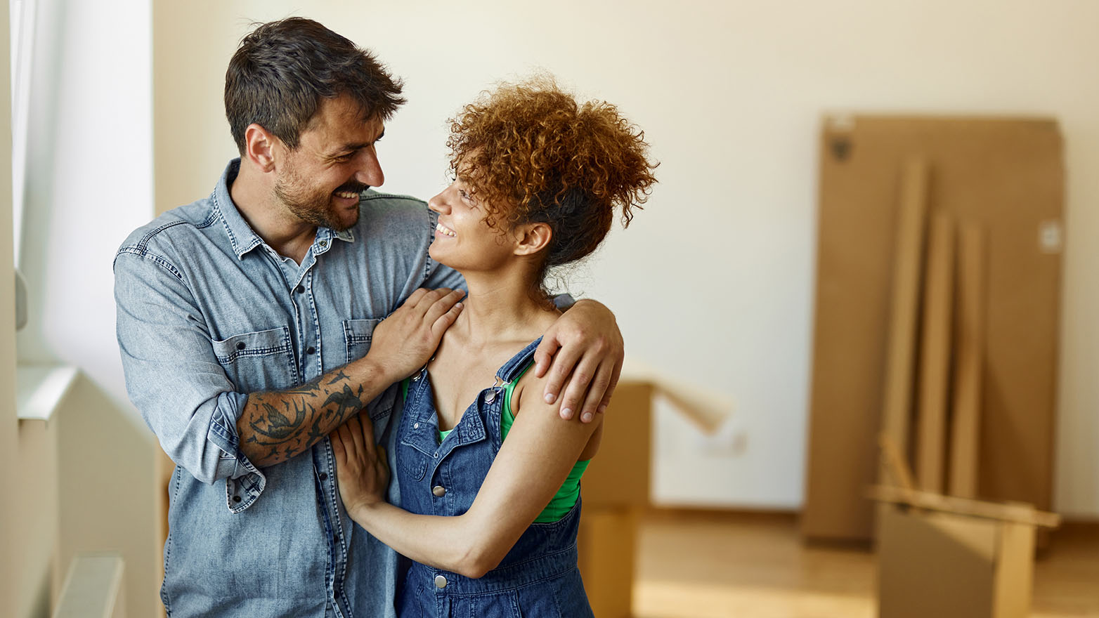 A young couple, bathed in the glow of a harshly bright room, stands amidst the remnants of a recent move. The man, with his dark hair and a beard meticulously kept, sports a tattoo on his left arm and a light blue denim shirt. His partner, a woman with a crown of curly hair, dons a green top and a denim dress. They are wrapped in each other's arms, beaming and lost in each other's gaze. The room, with its sparse furnishings, is a blank canvas of white walls and white moldings.