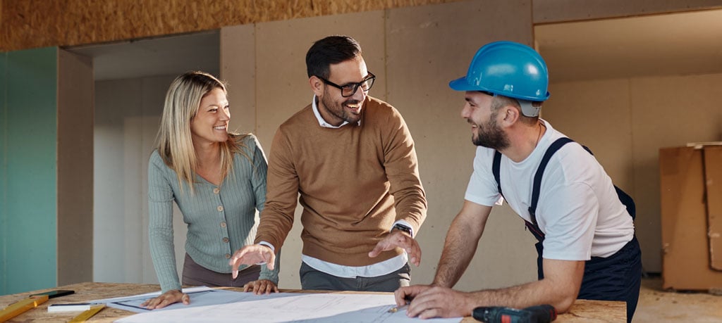 A smiling couple and a construction worker wearing a blue hard hat review blueprints on a wooden table in a partially constructed building. The couple looks engaged and pleased as the worker explains the plans, creating a sense of collaboration and excitement about the construction project.