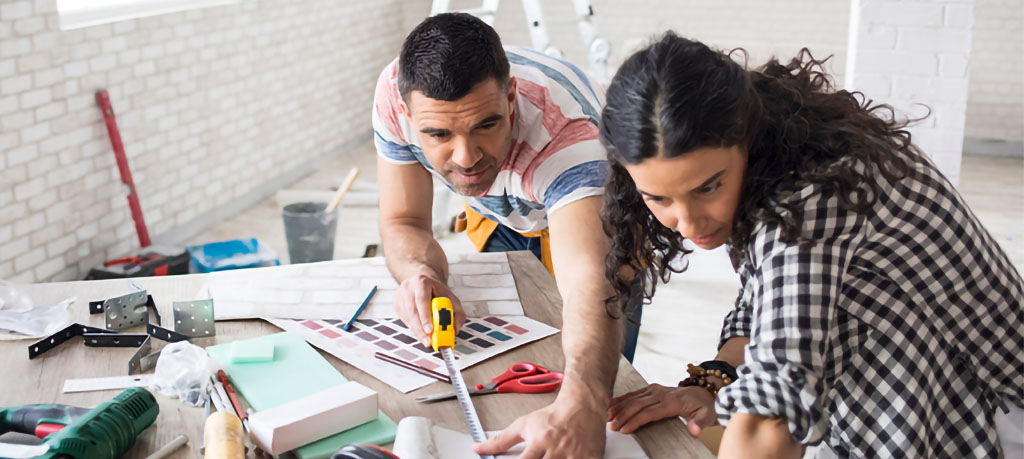 A couple is planning a home renovation. They are leaning over a table with a tape measure, paint swatches, and tools. They appear to be discussing the project, possibly measuring materials or selecting colors. The room behind them has a partially finished look, suggesting they are in the midst of the renovation process.