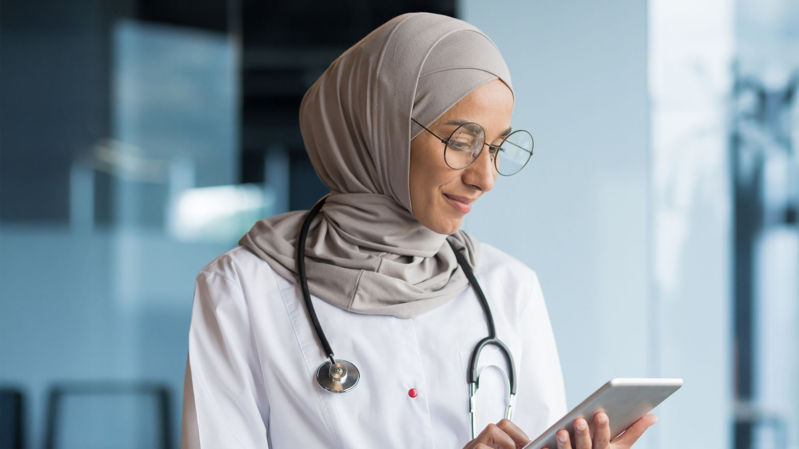 Medium shot of a female doctor with a light grey hijab and round, wire-rim glasses, looking down at a tablet. She is wearing a white lab coat with a stethoscope around her neck. She looks gentle and attentive. The background is a soft, blurred blue, suggesting a modern office or hospital setting.