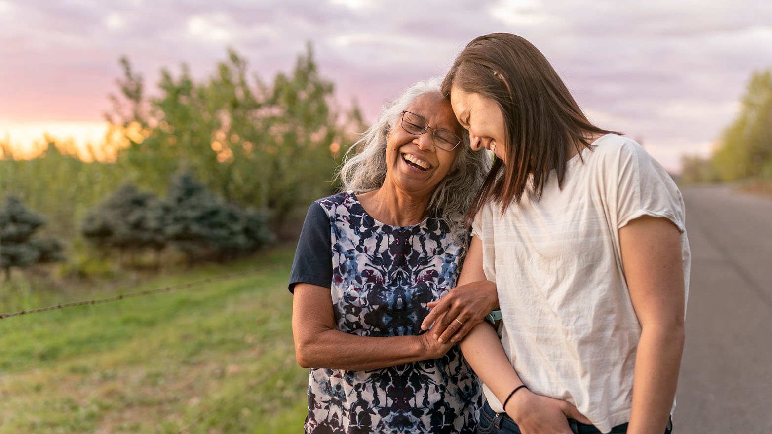 Two women, an older adult with silver hair and glasses and a younger woman, share a tender moment. They stand together outside, heads gently leaning against each other, both smiling. The older woman's face is tilted back in laughter. A natural background includes trees, a barbed wire fence, and a vibrant sky with colors of the sunset.