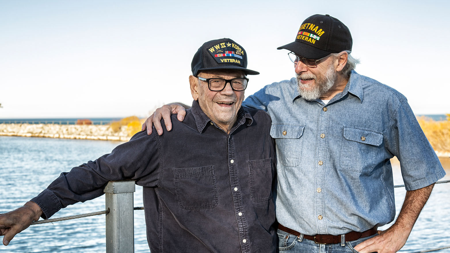Two distinguished veterans, brothers-in-arms, share a moment of camaraderie on a peaceful pier. To the left, a gentleman of the greatest generation, his cap a testament to his service in WWII and Korea, a warm smile gracing his face. To the right, a Vietnam Veteran, his cap a symbol of a different era, yet the bond of service unites them. Both men, in their own way, embody the spirit of duty and honor.
