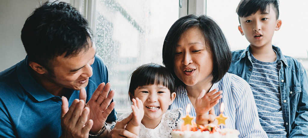 An Asian family of four is in the midst of a birthday celebration. A young girl is beaming at the camera, the star-shaped candles on her cake aglow, as her parents applaud and a boy stands behind them.
