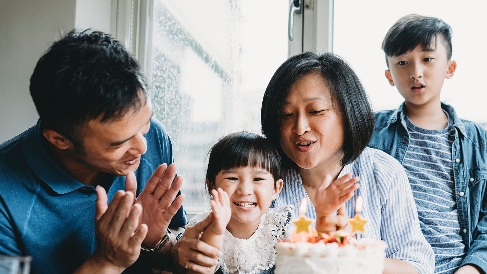 An Asian family of four is in the midst of a birthday celebration. A young girl is beaming at the camera, the star-shaped candles on her cake aglow, as her parents applaud and a boy stands behind them.