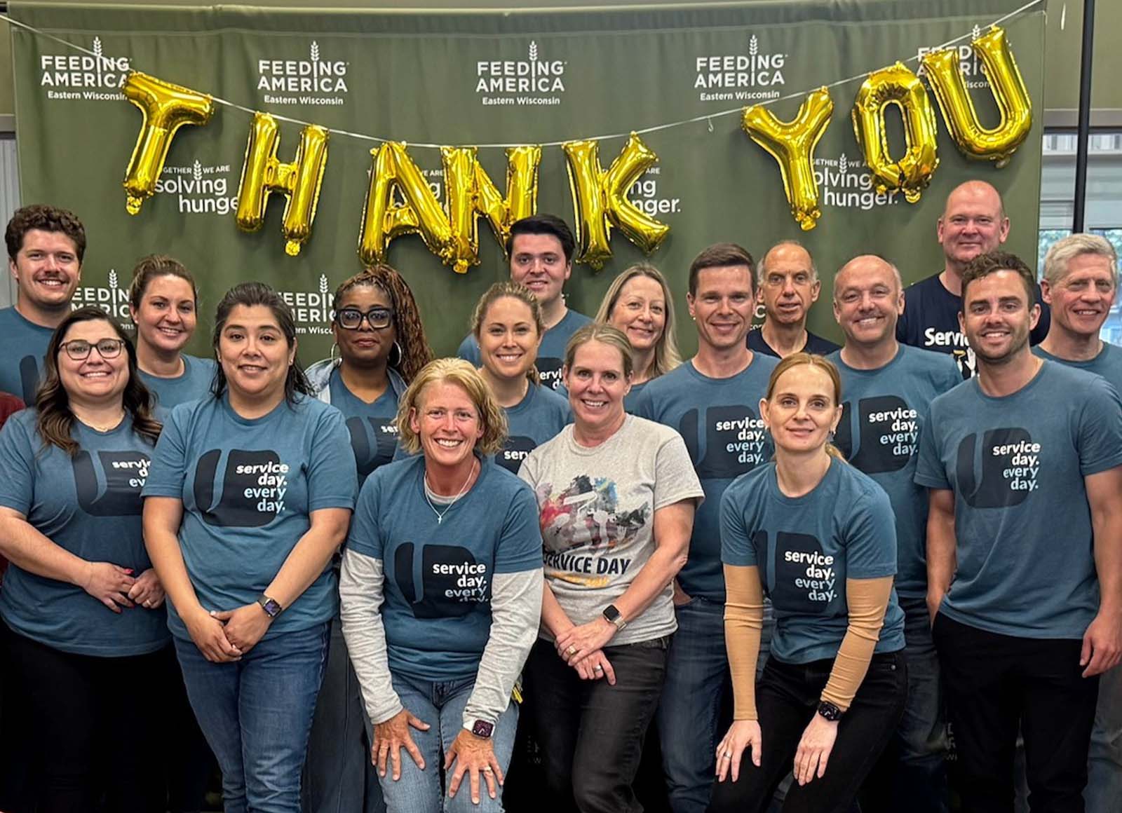 A group of about 20 people, mostly wearing blue "Service day. every day." t-shirts, stand together in front of a "Feeding America Eastern Wisconsin" banner. The banner is decorated with gold balloon letters that read "Thank You."