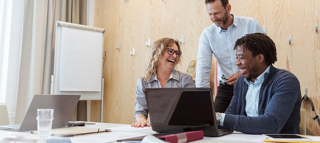 Three business colleagues, smiling, collaborate around a laptop.