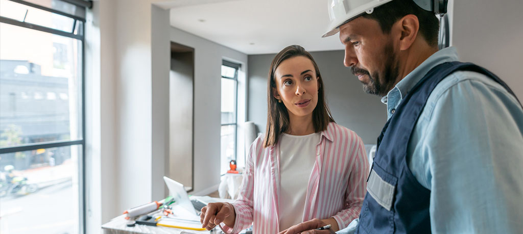 A woman in a pink and white striped shirt is in deep conversation with a construction worker, who's all business in his hardhat, with a clipboard and pencil at the ready. The room behind them is a work in progress.