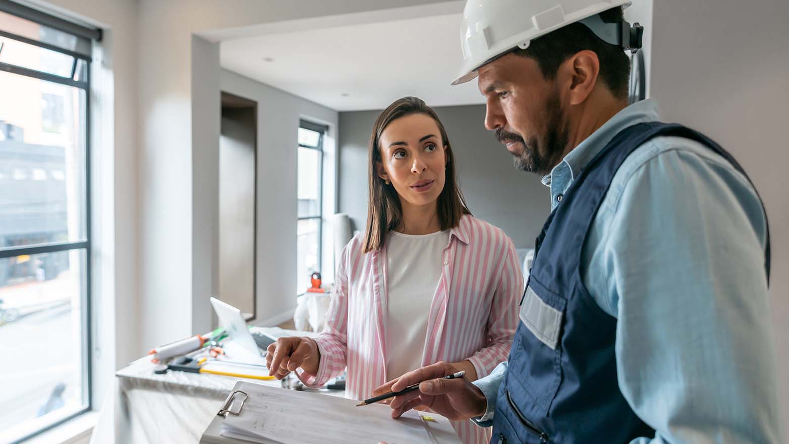 A woman in a pink and white striped shirt is in deep conversation with a construction worker, who's all business in his hardhat, with a clipboard and pencil at the ready. The room behind them is a work in progress.