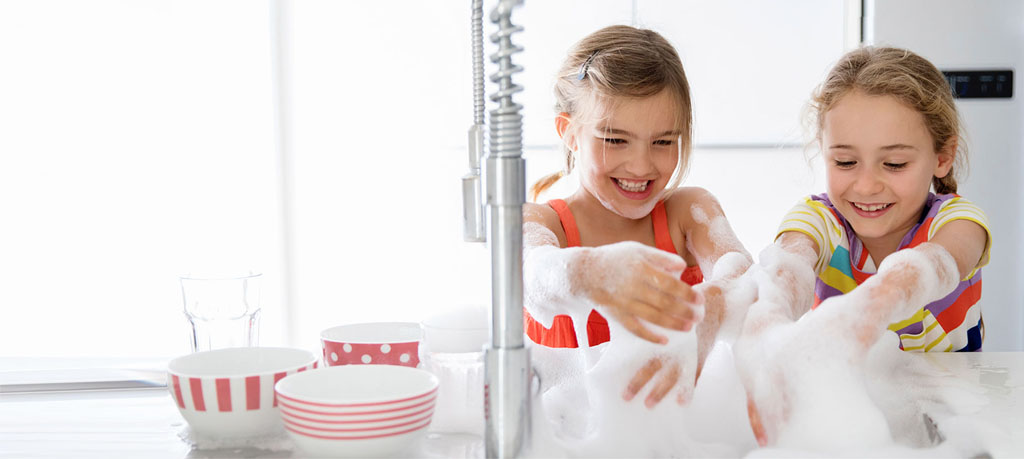 Two young girls, around the age of 7, are positioned side by side at a white sink. They are enthusiastically playing with a mountain of white sudsy bubbles. The girl on the left has her hair pulled back from her face with a small hair clip, and she is wearing an orange tank top. She's looking directly at the viewer and smiling broadly, her teeth showing. The girl on the right has her hair pulled back into braids and is wearing a colorful striped shirt with yellow, white, purple, red, and blue stripes. She's looking down at the bubbles, also with a smile on her face. The background is bright and clean, with a white refrigerator visible behind them. A chrome kitchen faucet is prominent on the left side of the image. The overall impression is one of fun, innocence, and joyful play.
