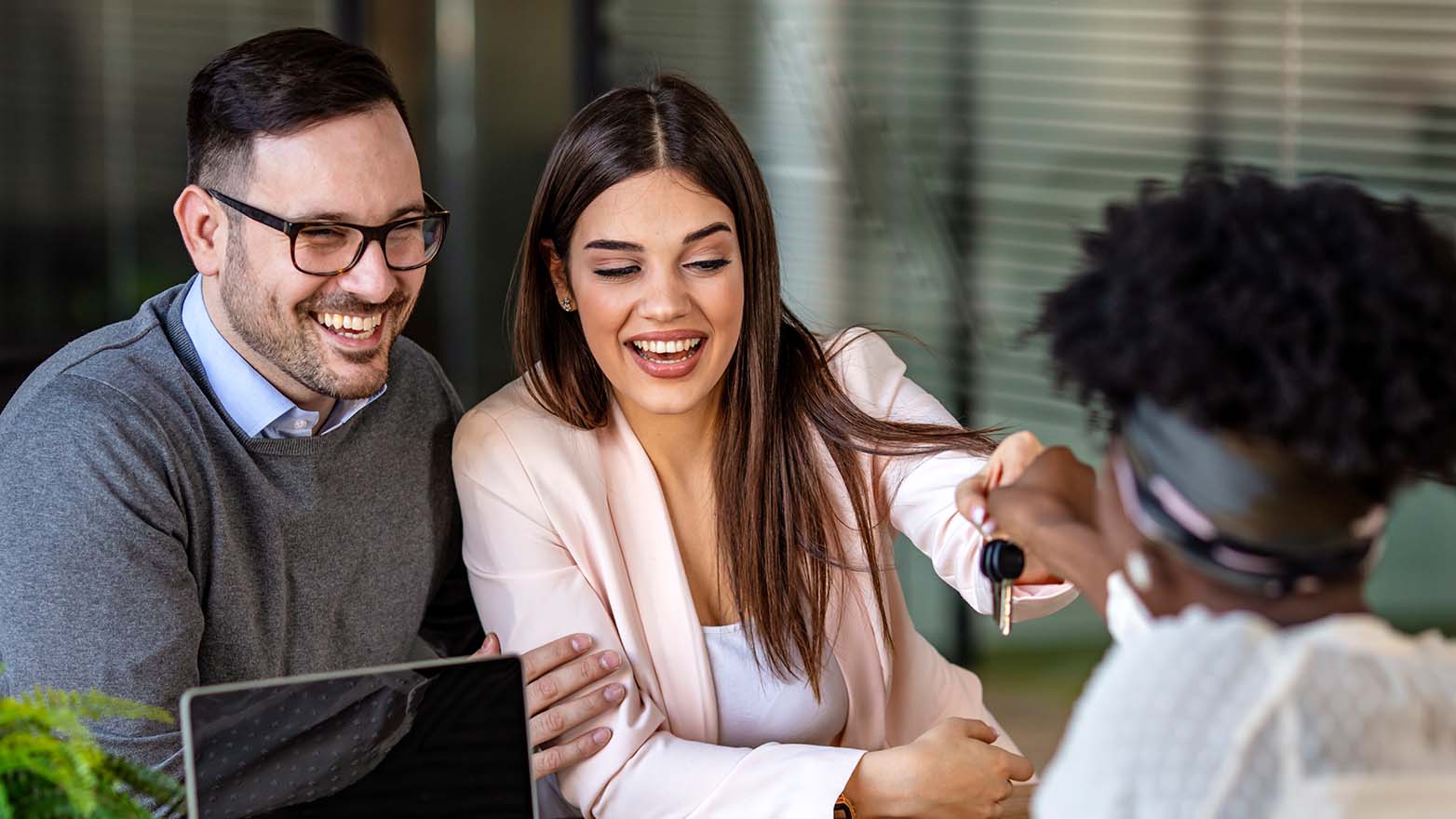 A couple, both smiling, are sitting down. The man, wearing a grey sweater and glasses, is to the left, and the woman, in a pink blazer, is to the right, looking down at the keys she's just been handed. There's a laptop on the table in front of them, and a plant in the corner. The sales representative, with an afro and a light-colored top, is handing over the keys.
