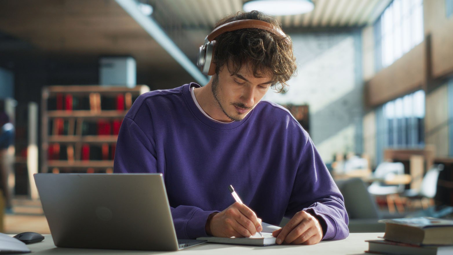 A young man with curly hair, wearing headphones, sits at a library desk, typing on a laptop while taking notes in a nearby notebook.