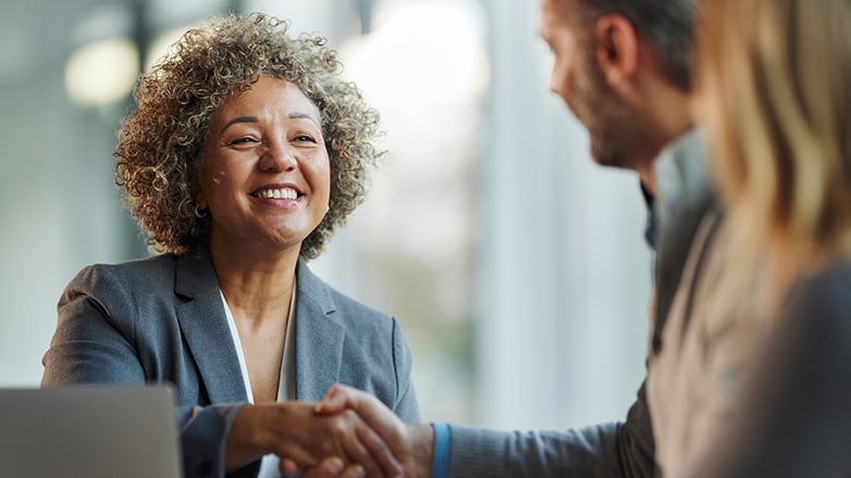 Two women, one with curly brown hair and a smile, and the other with blonde hair, stand on either side of a man in a gray suit, as they exchange a handshake.