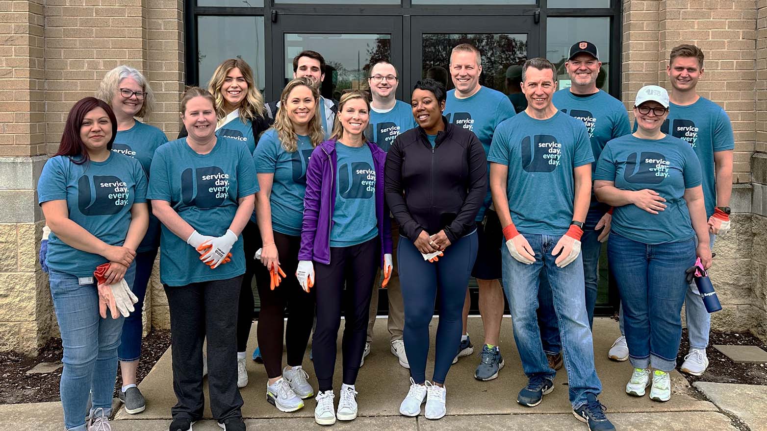 Three people, all smiling and wearing matching blue t-shirts and orange safety vests, stand together in front of stacked boxes in a warehouse setting.