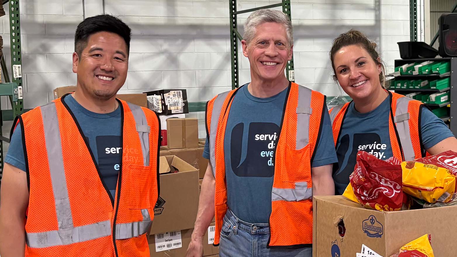 A diverse group of 14 people, varying in age, gender, and ethnicity, stand together outside a building. They wear matching teal shirts with the phrase "service day. every day." and many have on work gloves. The building's exterior features light brick and dark windows.