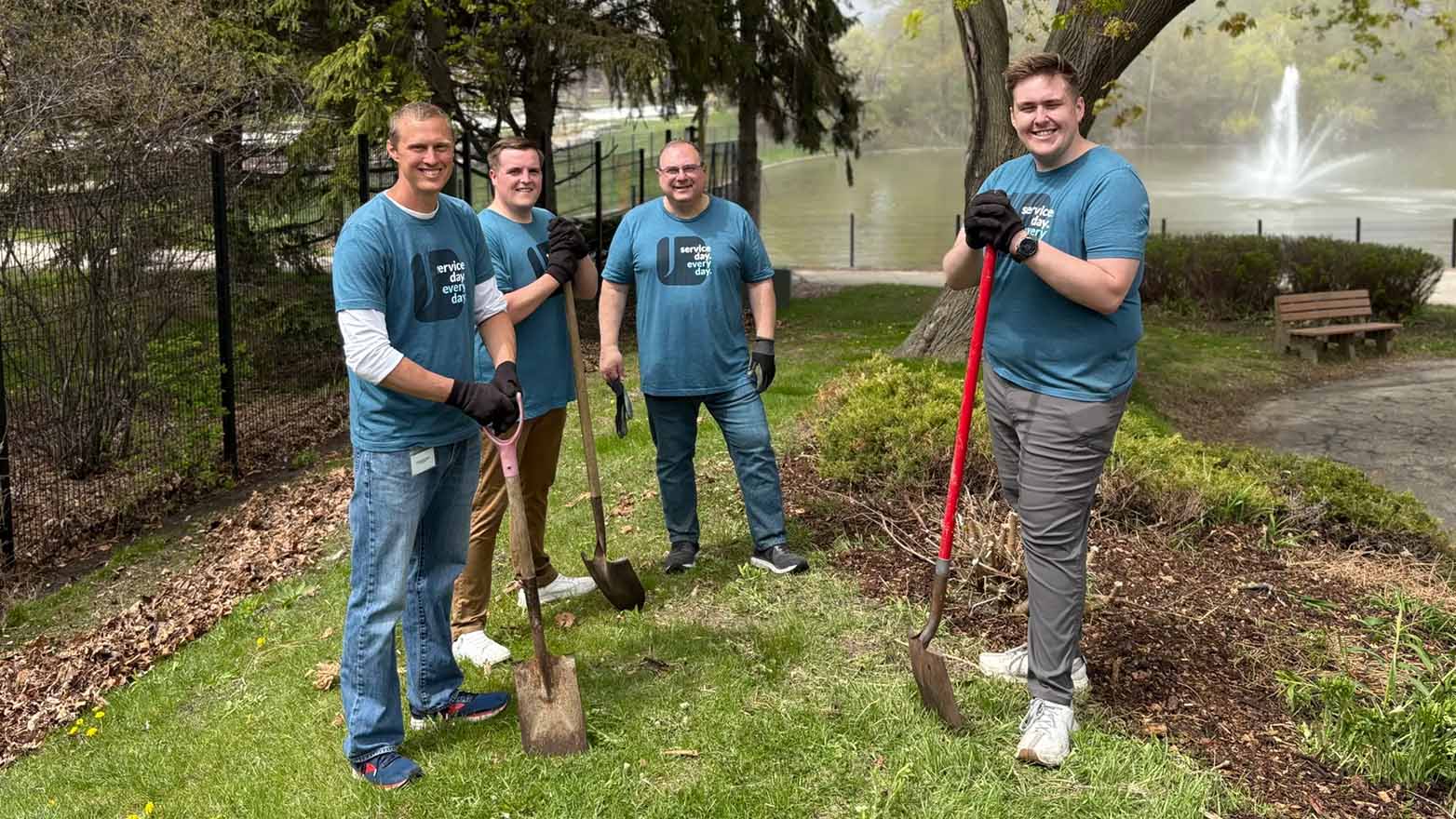 Four people wearing matching shirts stand together, each holding a shovel, in a park setting.