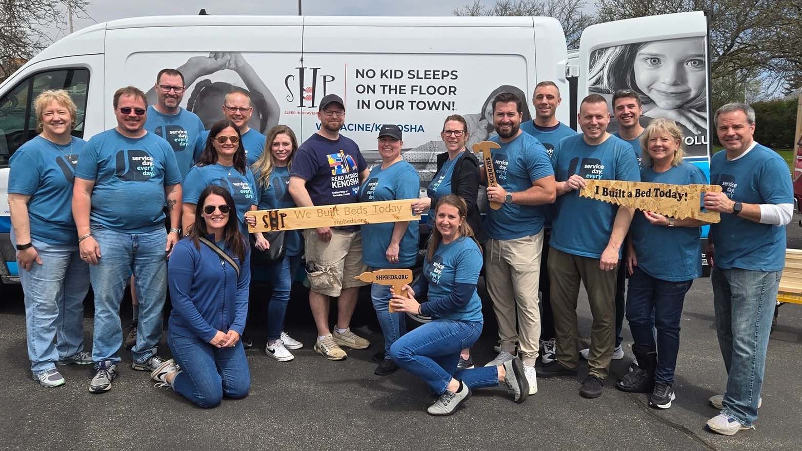 A group of people stand together, smiling, holding up handmade signs in front of a van. One sign reads, "SHP We Built Beds Today" and another says, "I Built a Bed Today!".