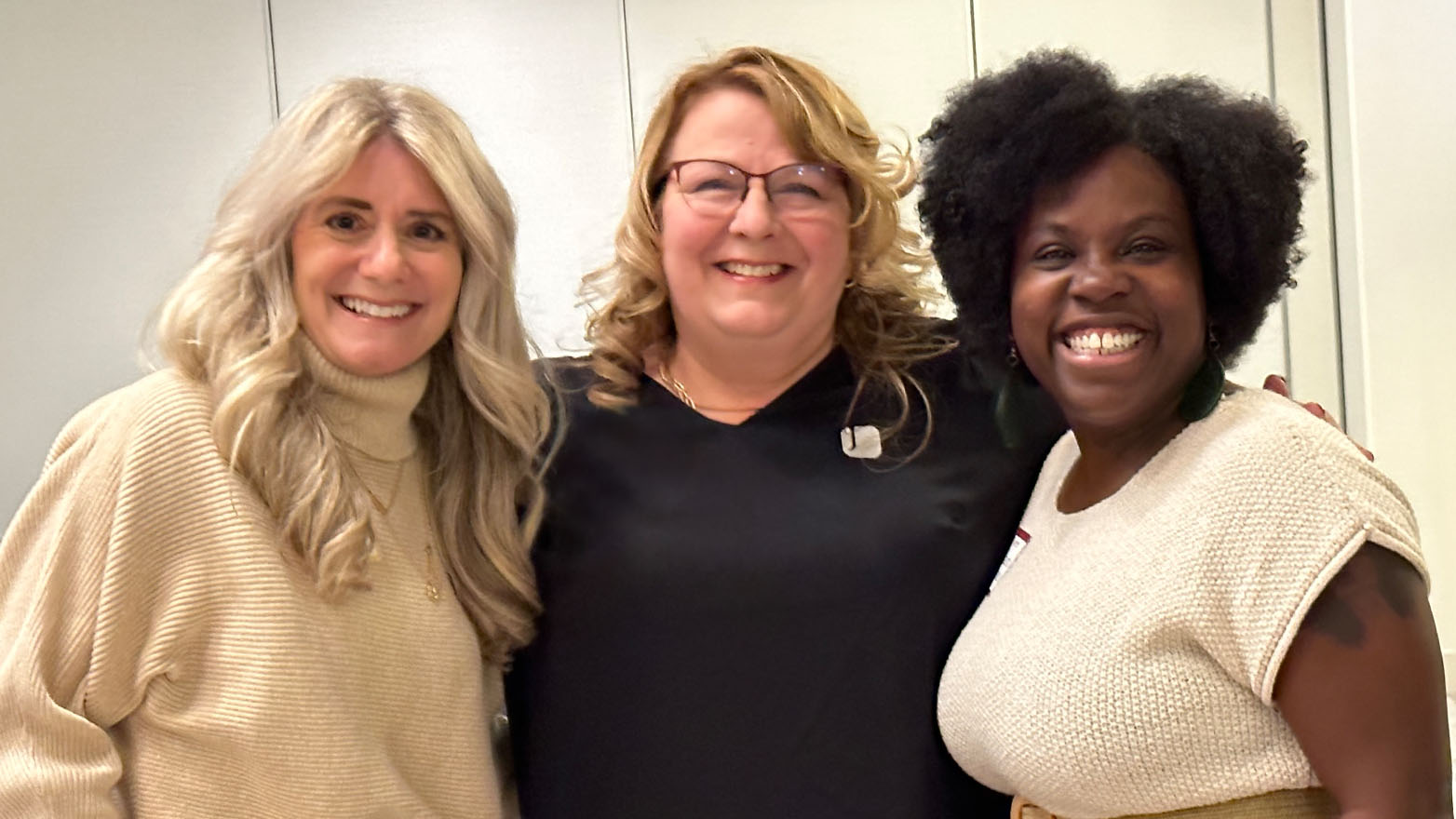 Three women stand together, smiling and facing the camera, their shoulders almost touching.