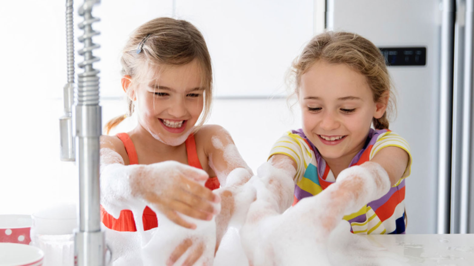 Two young girls, around the age of 7, are positioned side by side at a white sink. They are enthusiastically playing with a mountain of white sudsy bubbles. The girl on the left has her hair pulled back from her face with a small hair clip, and she is wearing an orange tank top. She's looking directly at the viewer and smiling broadly, her teeth showing. The girl on the right has her hair pulled back into braids and is wearing a colorful striped shirt with yellow, white, purple, red, and blue stripes. She's looking down at the bubbles, also with a smile on her face. The background is bright and clean, with a white refrigerator visible behind them. A chrome kitchen faucet is prominent on the left side of the image. The overall impression is one of fun, innocence, and joyful play.