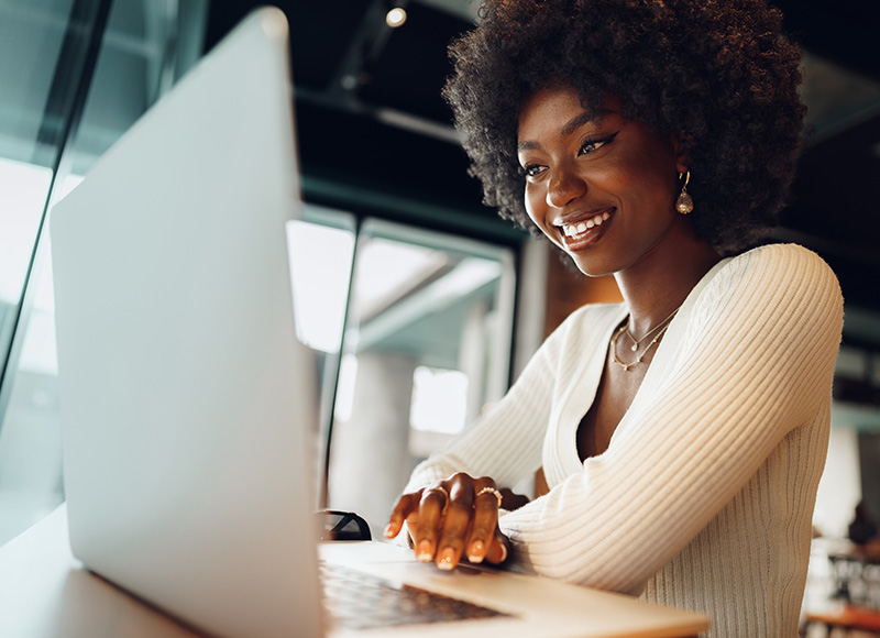 A Black woman with a large afro smiles as she uses a laptop computer in a cafe.