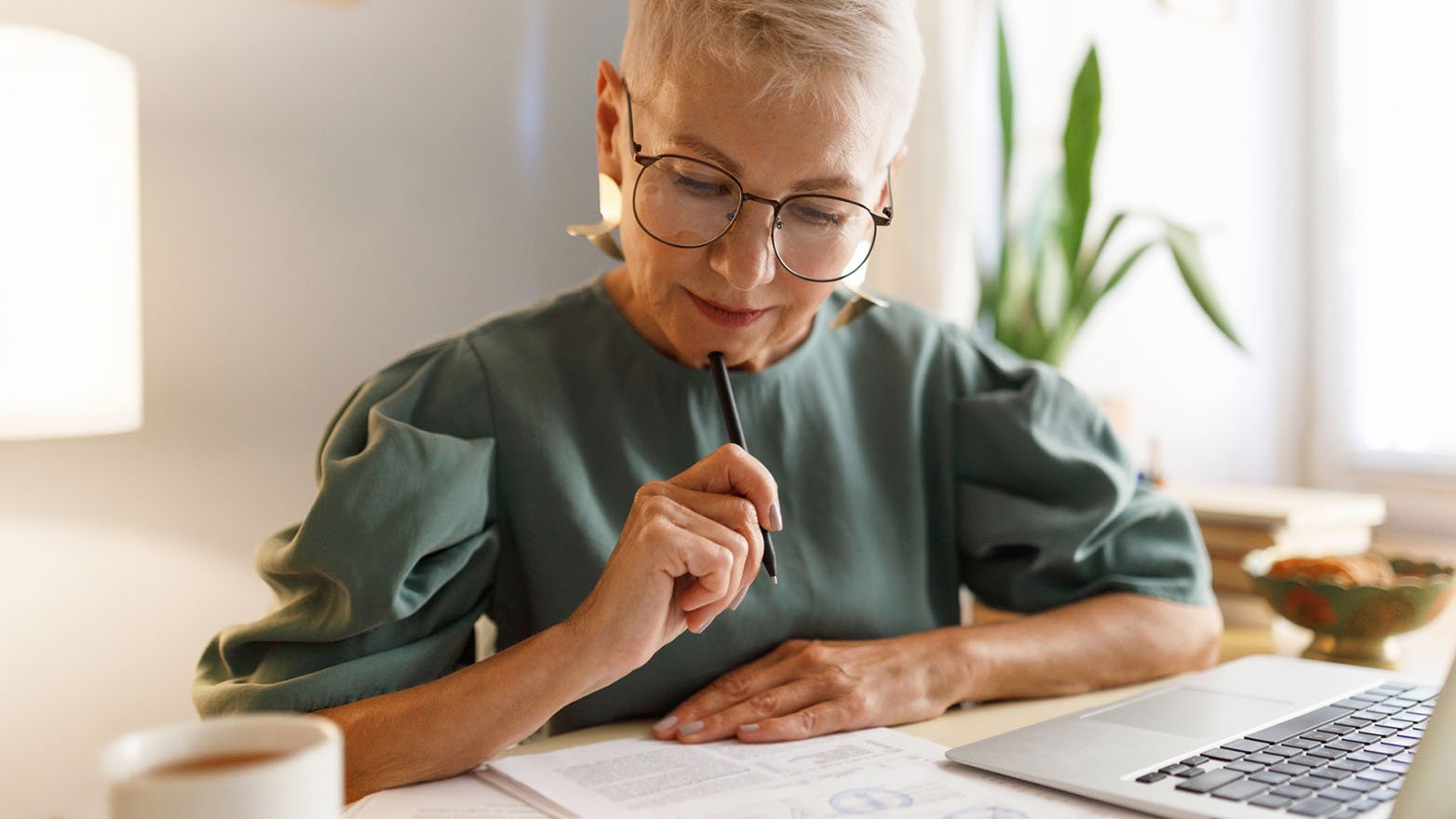 A middle-aged woman with glasses sits at a desk, deeply in thought, with her chin resting on a pen. A laptop and documents are spread out before her.