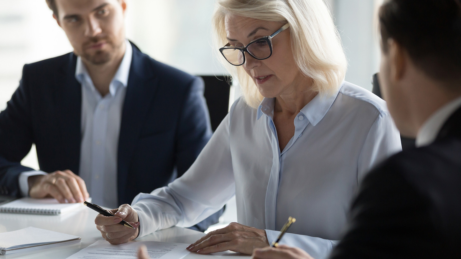A mature woman with blonde hair and glasses is signing a document at a table, while two other business people in suits observe. Pens and papers are visible.