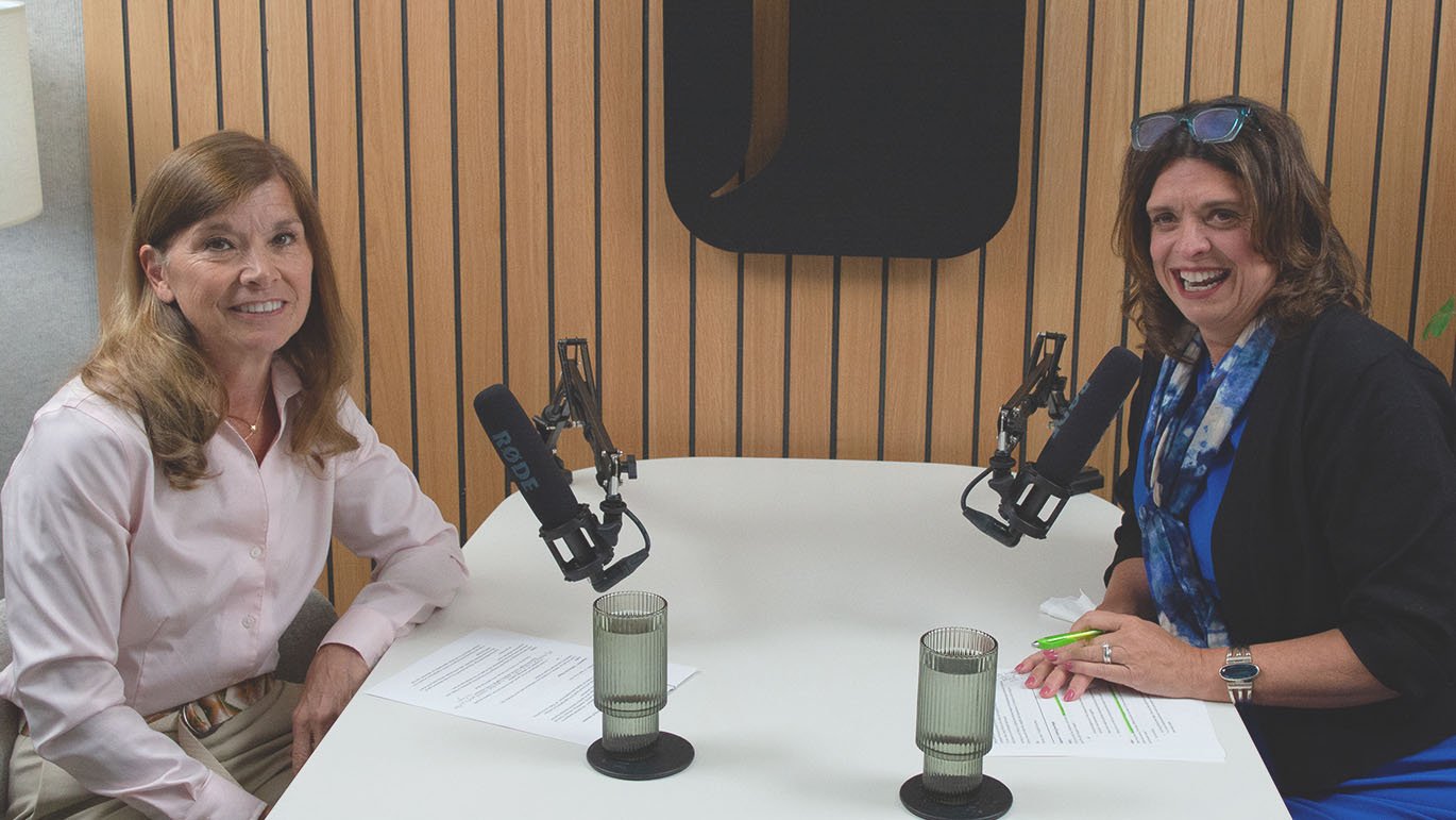 Two women sit at a white table, each positioned at a microphone, preparing to record a podcast.