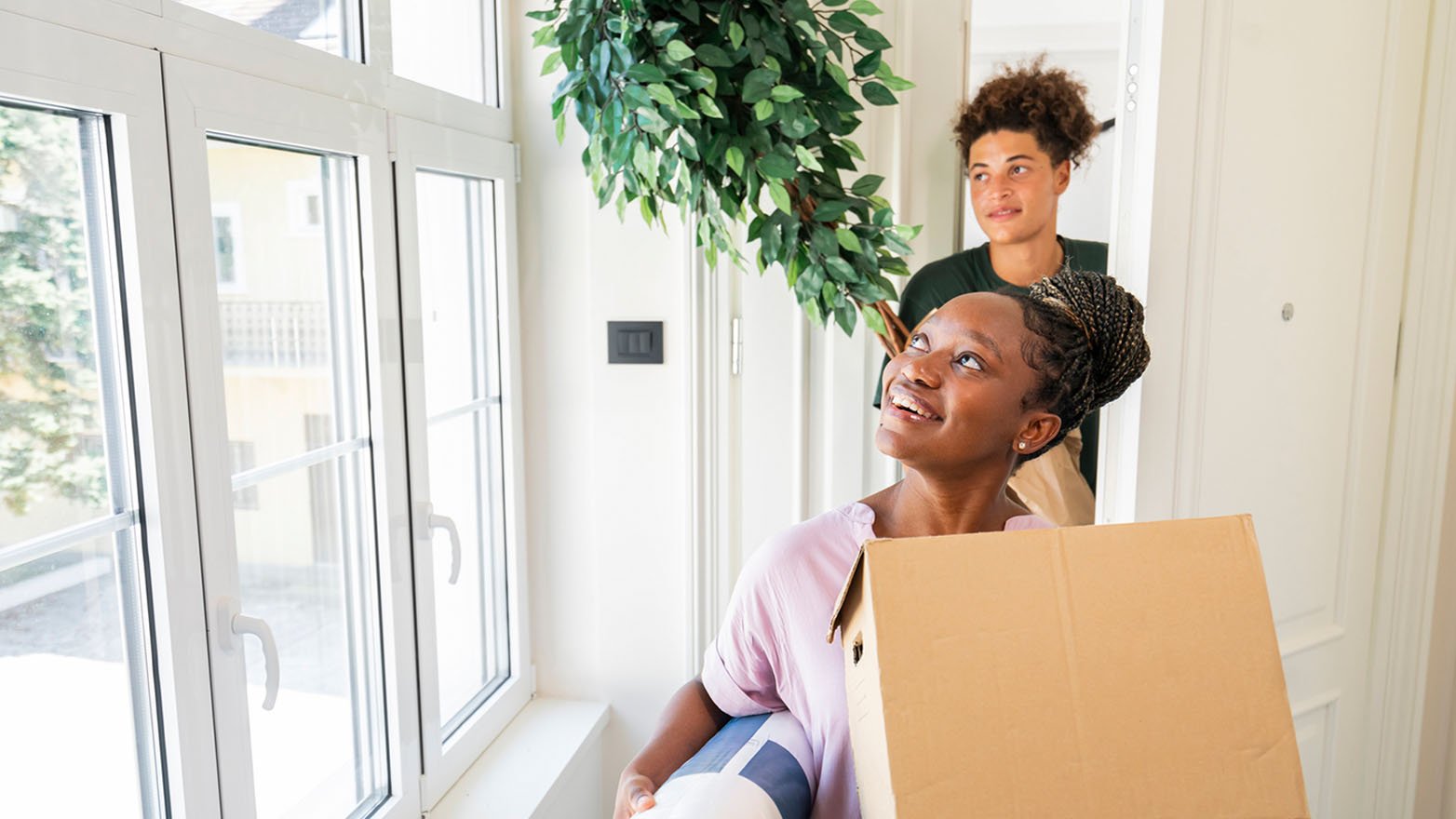 A young Black woman, with a cardboard box and rolled-up rug in hand, gazes up with a smile as she steps into a sunlit room. Behind her, a Black man assists with a sizable potted plant. The scene suggests a new home in the making.