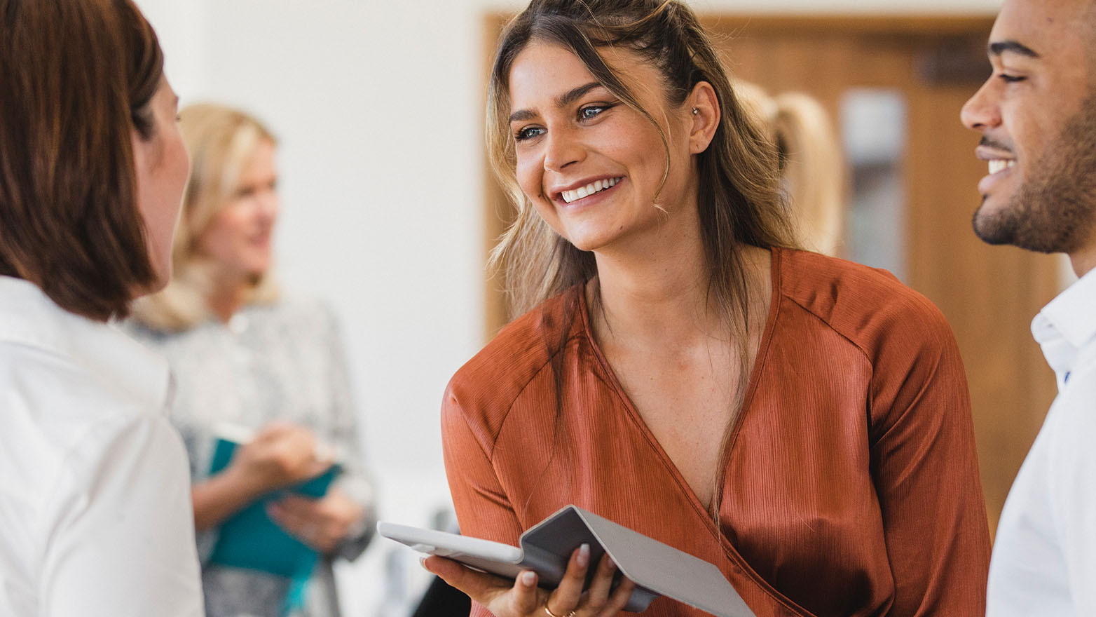 A group of professionals with diverse backgrounds are gathered in a brightly lit office space, engaged in what appears to be a lively discussion or meeting. The focal point is a young woman with light brown hair, a few strands pulled back, who is smiling and looking at someone off-camera. She is wearing a rust-colored blouse and holding a tablet. To her right, a man with dark hair and a well-groomed beard is also smiling, contributing to the convivial atmosphere. In the background, other individuals are partially visible, one of whom is holding a blue notebook, indicating an informal, collaborative setting. The lighting is soft and diffused, emphasizing the group's friendly and approachable nature.