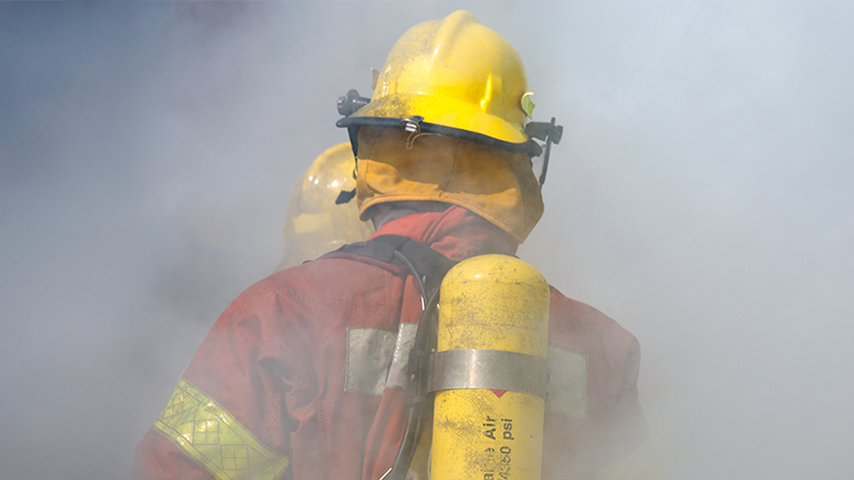 A firefighter's back is visible, wearing a yellow helmet, red turnout gear with yellow reflective strips, and a yellow air tank strapped to their back. The scene is filled with thick smoke, suggesting an active fire. Another firefighter's helmet is partially visible in the background.