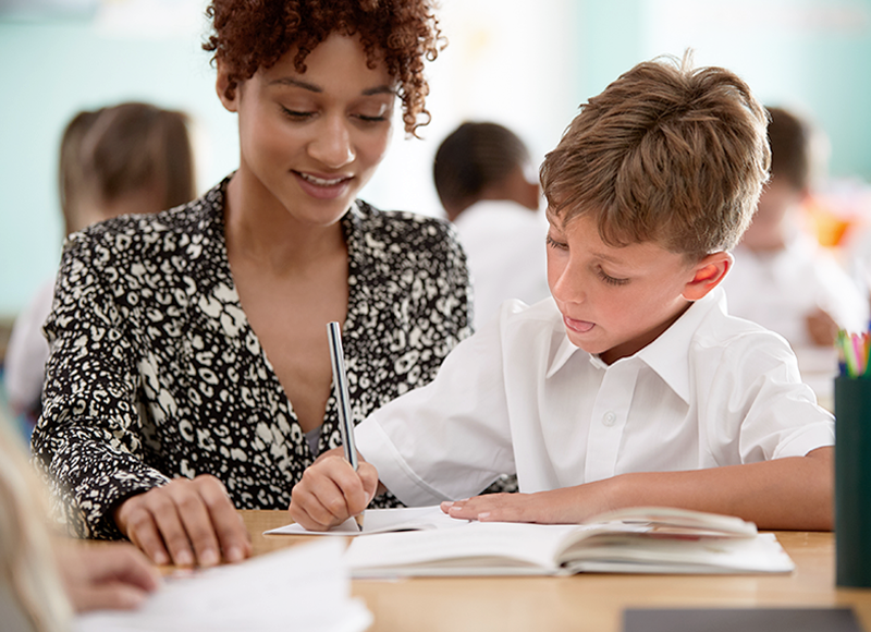 A young female teacher with curly hair, wearing a black and white patterned blouse, leans over to help a male elementary school student in a white shirt write in a notebook. Other students are visible in the blurred background of a classroom.